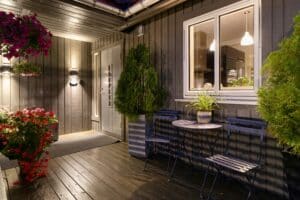 Night view of a covered wooden porch with black siding, a glass door, a white-framed window, and blue metal seating; pink flowering hanging basket and potted greenery add color under warm lights on the wall.