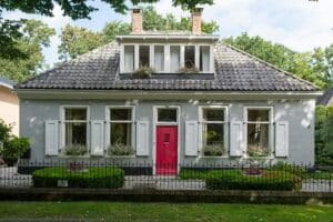 Charming gray cottage-style home with white shutters, a red front door, manicured landscaping, and a tiled roof surrounded by mature trees.