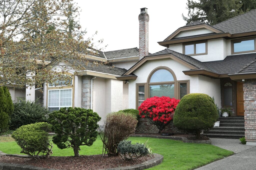 Front view of a suburban house with a brick chimney, curved arched window, and manicured bushes in a green yard.