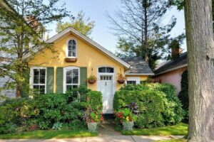 Charming yellow cottage-style home with mature landscaping, flowering plants, and a welcoming front entrance surrounded by trees.