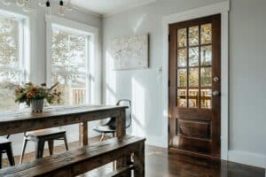 Bright dining room with large windows, rustic wood table, and glass-paneled front door, showcasing natural light and inviting home appeal.